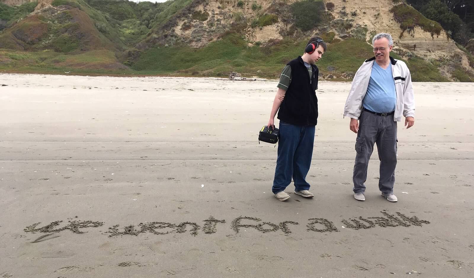 A student and a teacher at the beach with the sentence 'We went for a walk' written in the sand in front of them